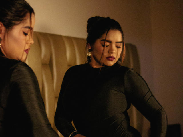 NinnaRose with closed eyes, wearing a black, long-sleeve dress, sits on a beige, tufted chair. Her dark hair is in a bun, wearing gold earrings. Boobs visible.