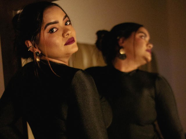 NinnaRose with dark hair in a bun, wearing a sheer black top, facing a mirror showing her profile. She has purple lipstick, gold earrings, and a serious expression.