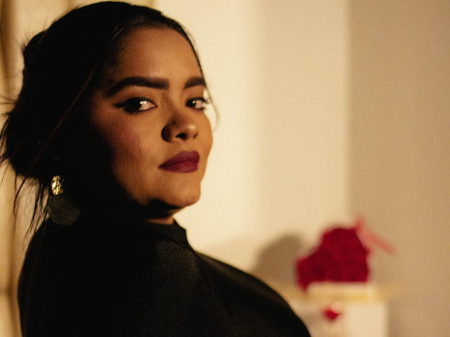 NinnaRose with dark hair, deep red lipstick, and black earrings, looking over her shoulder with a subtle smirk, wearing a black top, in a softly lit, warm-toned photo.
