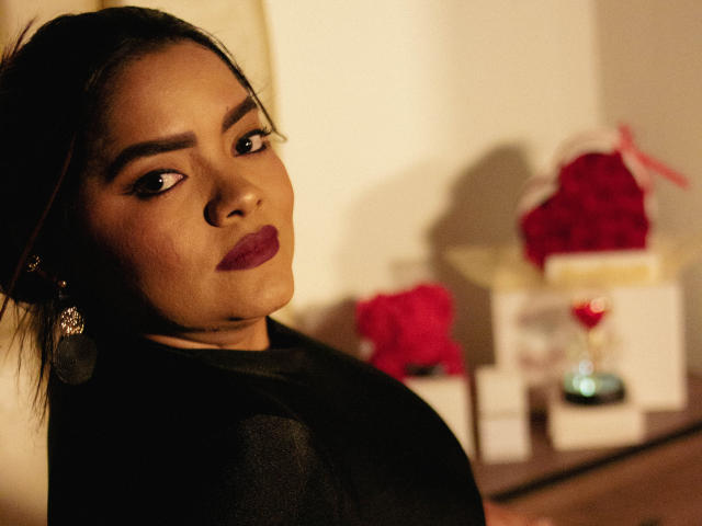 NinnaRose with dark hair, red lipstick, black top, and hoop earrings, looking over her shoulder, her face and upper body in focus, blurred red and white objects in background.