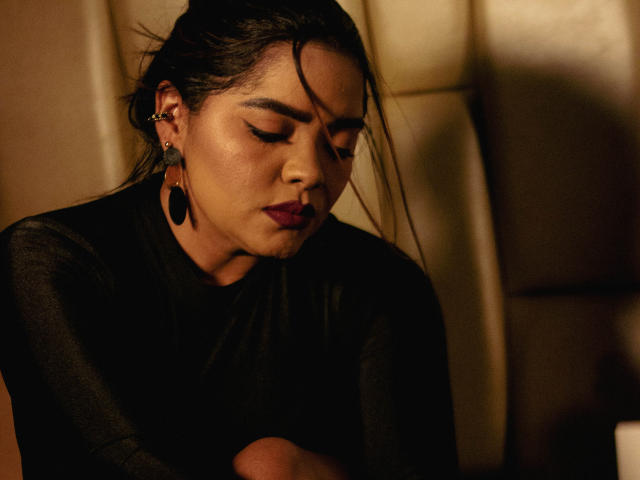 NinnaRose with dark hair, wearing a black sheer top, looking down with serious expression, purple lipstick, and large black earrings, sitting in dim, warm lighting.