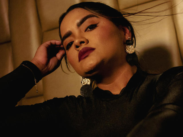 NinnaRose with dark makeup, wearing black earrings and shirt, hand in hair, looking down with a serious expression, warm lighting, soft shadows, slight smile.