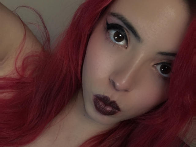 Rosalinnes with red hair, dark eyeliner, and matte purple lipstick, pouting her plump, glossy purple lips, looking at the camera with her large, brown eyes.
