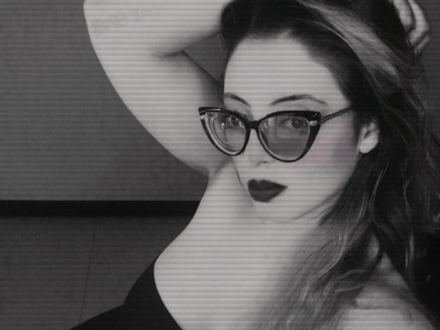 Black-and-white photo of Rosalinnes with glasses, dark lipstick, and shoulder exposed, arm raised, long hair, looking at camera, showing her tits.