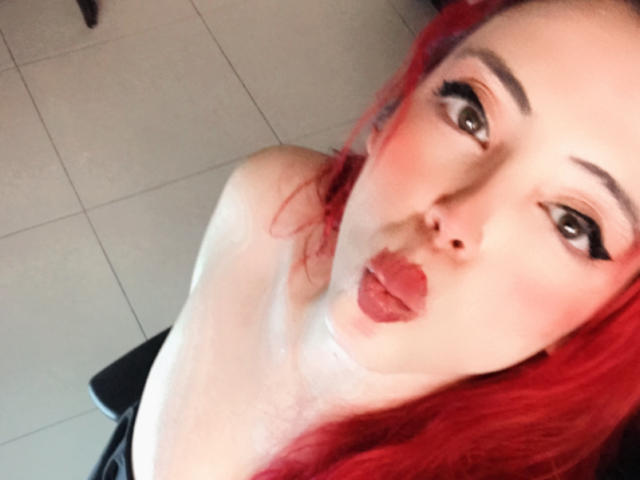 Rosalinnes with red hair, pouty lips, and heavy eyeliner, showing a drop of white liquid on her neck and chest. Boobs partially visible. Rosalines' face is angled slightly downward.