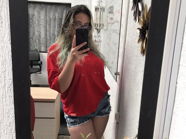 Rosalinnes with long green-tipped hair, glasses, red shirt, and denim shorts, taking a mirror selfie, showing her ass and tits, in a room with a window.