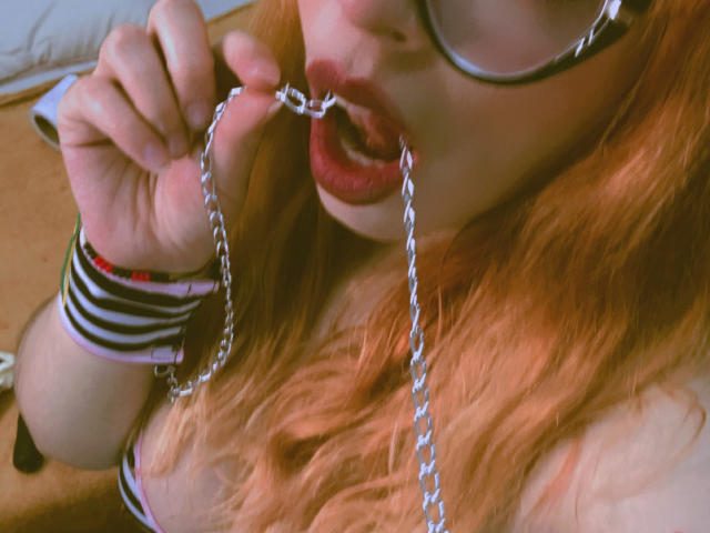 Rosalinnes with red hair, wearing glasses, biting a metal chain, lips open, showing her tits, striped wristbands, in a sexy, provocative pose.