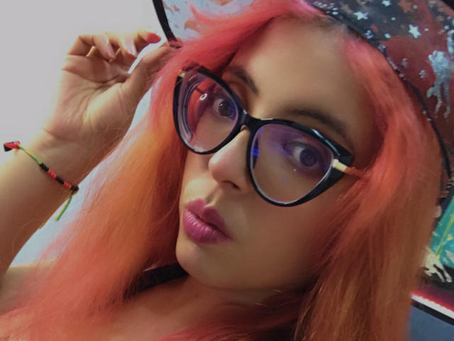 Rosalinnes with pink hair, purple lips, black glasses, and a star-patterned hat, holding her hair. She's wearing a colorful bracelet. Her eyes are purple.