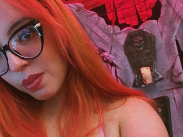 Rosalinnes with red hair, glasses, and pink lipstick, showing off her tits, a lit candle, and a statue in the background. Rosalinnes looks cute and sexy.