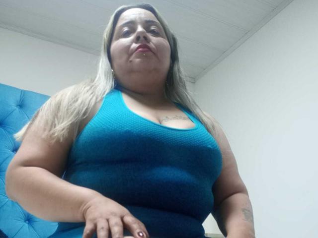 KyaraStone, a plus-sized woman with blonde hair, wearing a tight blue tank top, showing her large tits and tattoos, looking down at the camera with a neutral expression.