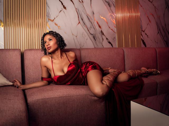 BriahLarson in a red satin dress, lying on a pink sofa with her tits showing, curly hair, gold strappy heels, looking to the side, sexy and seductive.