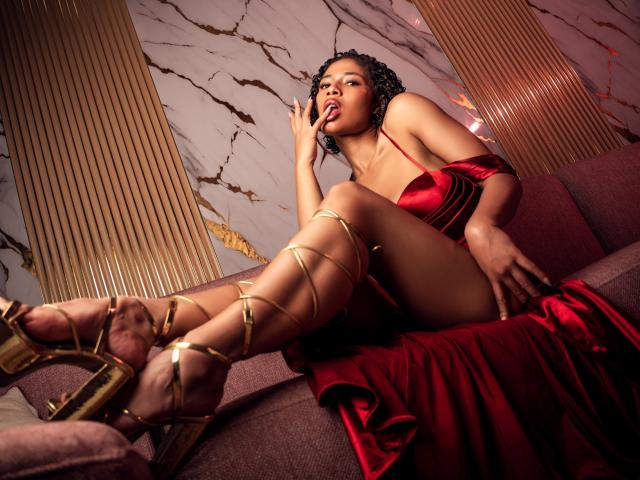 BriahLarson in red satin dress, gold strappy heels, sitting on purple couch, legs crossed, hand near mouth, curly black hair, sexy, brown skin, looking up, seductive pose.