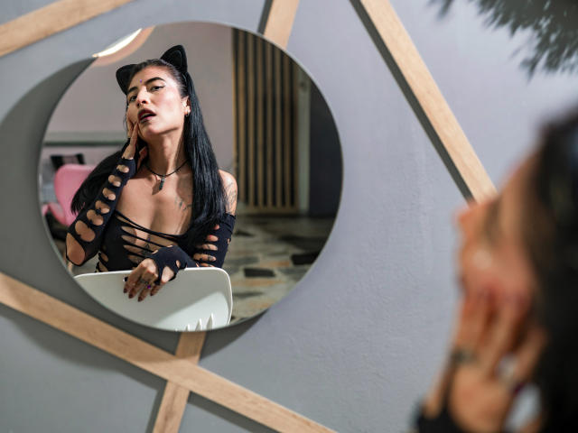 SexyRedLady with black cat ears, long black hair, and cut-out black top, looking in mirror, showing her tits, wearing fingerless gloves, in a modern room. Reflection visible.