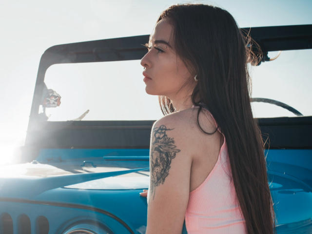 ArtemisSolo with long brown hair, pink tank top, tattoo on right shoulder, standing in front of a blue jeep, sunlight from left, profile view, soft light, relaxed expression.