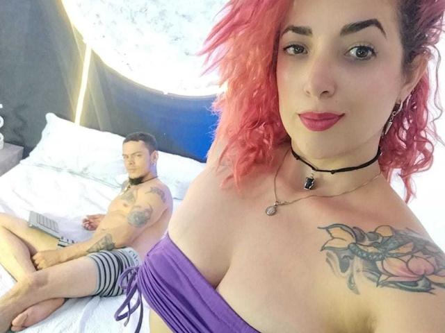 AbbyAndVincent with pink curly hair, big tits, tattoo on chest, wearing purple top, black choker, standing, smiling, looking at camera. Vincent, shirtless, tattooed, striped shorts, sitting on bed.