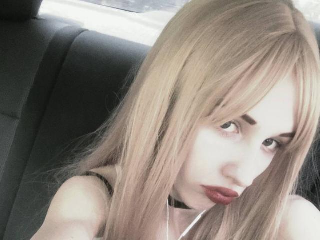 EnchantedFay has blonde hair, red lipstick, and white earbuds, with a black choker, looking over her shoulder in a car. Her tits are partially visible.