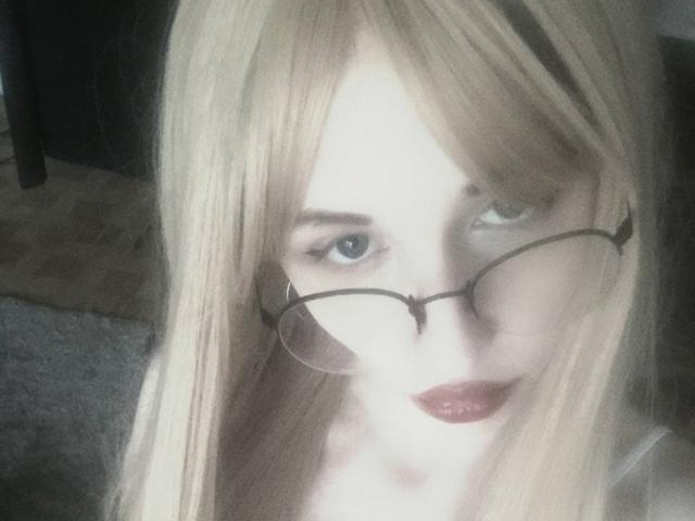 EnchantedFay with long blonde hair, glasses, and dark red lipstick, has pale skin and blue eyes, looking slightly to the side with a subtle, almost shy smile.