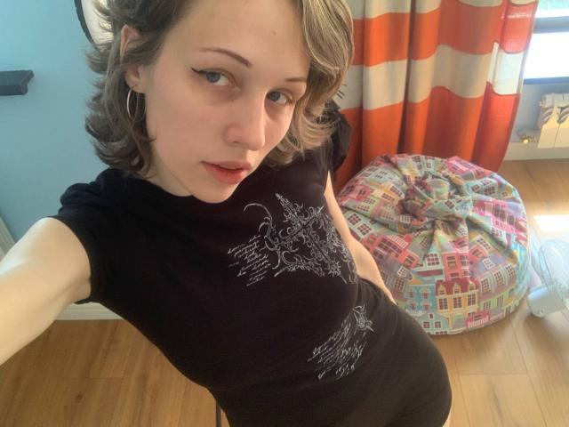 EnchantedFay with short brown hair, black t-shirt, and hoop earrings, taking a selfie. She's looking at the camera with a slight smile, her tits barely visible.