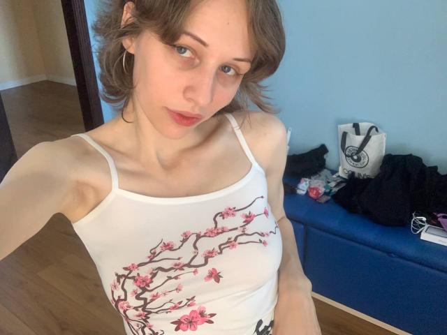 EnchantedFay with short brown hair, green eyes, and hoop earrings, wearing a white tank top with pink cherry branch design, showing small tits and slim arms, looking at camera.