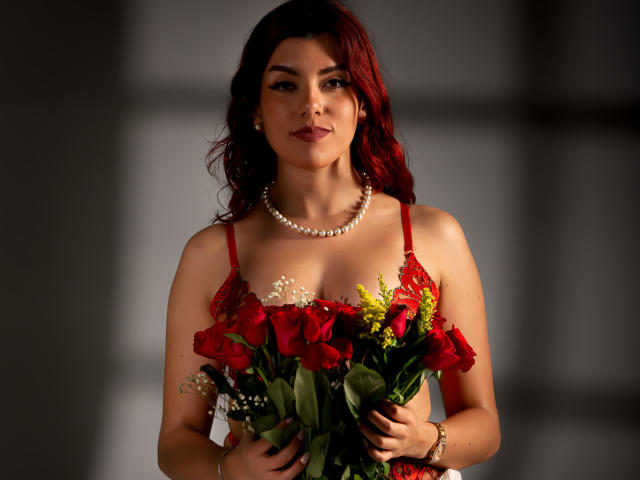 ZoeeMar with red hair, holding a bouquet of red roses and yellow flowers, wearing a red lace dress and pearl necklace, looking at the camera with a slight smile.