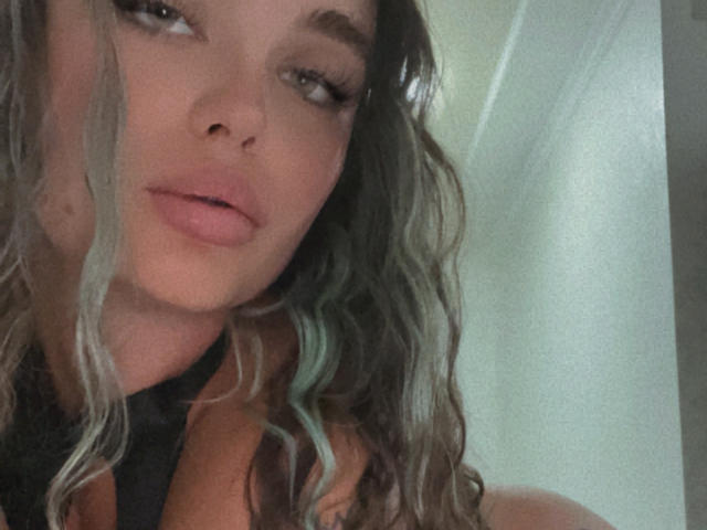 StarStorm has wavy, grayish-brown hair with blue tips, full pink lips, and a slightly pouty expression. She's wearing a black top, and her eyes are half-open.