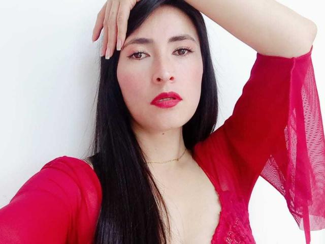 JainaRogers with long black hair, red lipstick, and red sheer top, hand on head, looking at camera, pale skin, small gold necklace, white background.