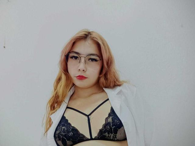 LiliithStorm with long pink hair, glasses, red lipstick, wearing an open white shirt and black lace bra with straps, looking straight at the camera. Boobs visible through lace.