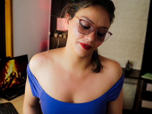 VeronicaSaienz with glasses, red lipstick, and a blue off-shoulder top, looking down with a slight smile, showing her bare shoulders and upper chest. Her tits are slightly visible.