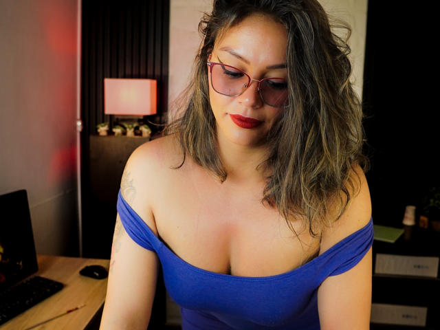 Veronica Saienz trägt roten Lippenstift, eine Brille und ein blaues Oberteil mit Schulterauschnitt, das ihre Brüste zeigt. Sie hat welliges, graubraunes Haar und ein Tattoo am linken Arm..