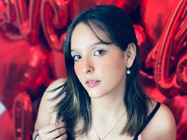 LanaKlyne, con cabello castaño, labios rosados, pechos pequeños, un top negro con tirantes, aretes y una sonrisa sutil, mirando ligeramente hacia un lado con una mano cerca de su hombro..