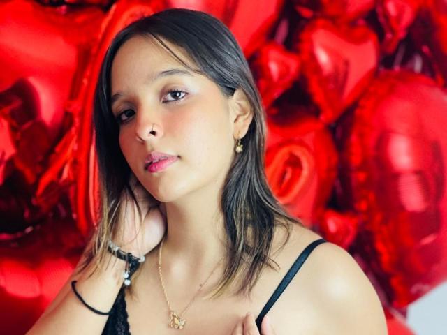LanaKlyne with light brown skin, small tits, brown eyes, and straight brown hair, wearing a black strap top, gold necklace, and earrings, against a red rose background.