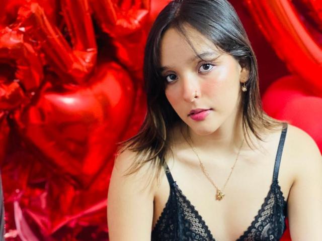 LanaKlyne with medium brown hair, wearing a black lace bra, gold necklace, and pink lipstick, looking slightly down with a neutral expression, red heart-shaped background.