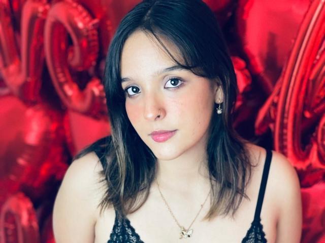 LanaKlyne with medium tits, wearing black lace, has light skin, brown hair, green eyes, pink lipstick, and a small nose ring, set against a red, shiny background.