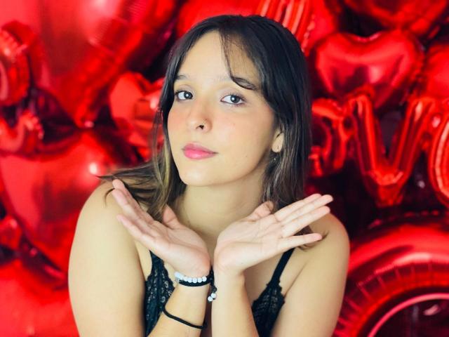 LanaKlyne with brown hair, pink lipstick, and black lace top, hands up, looking cute, wearing a black bracelet, surrounded by red heart-shaped balloons.