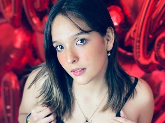 LanaKlyne with shoulder-length brown hair, wearing a black top, holding straps, looking slightly to the side, pink lips, small earrings, and a necklace, with a red background.