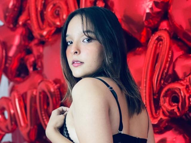LanaKlyne with brown hair, black bra, looking over shoulder, red shiny balloon background, holding bra strap, slightly parted pink lips, soft light, subtle makeup, sexy, confident.