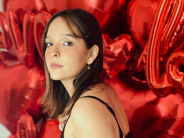 LanaKlyne with medium brown hair, black strap dress, small gold earrings, looking over shoulder, red foil balloons in background, subtle makeup, slight smile, soft lighting, medium shot.