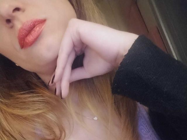 DonnaAngely's face, with glossy red lipstick, light skin, and blonde hair, touching her chin with a black-sleeved hand, wearing a purple top and a delicate necklace.