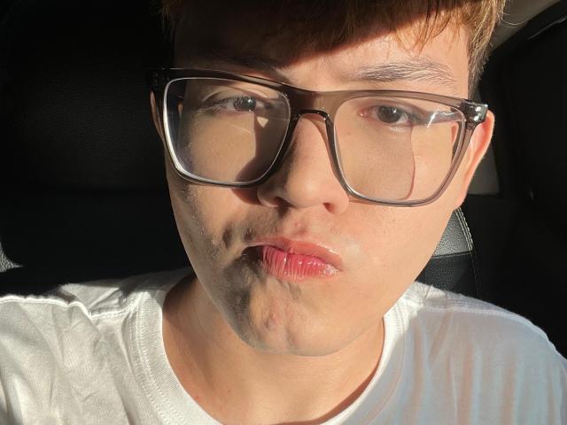 ErosDelRey with puffy, shiny lips, wearing large black-rimmed glasses, white shirt, and short brown hair, sitting in sunlit car. Reflections on glasses.