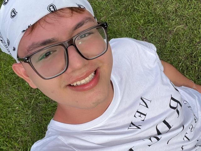 ErosDelRey with glasses, white bandana, and white shirt, lying on grass, smiling with visible teeth, short brown hair, and slight facial stubble, looking up at camera.
