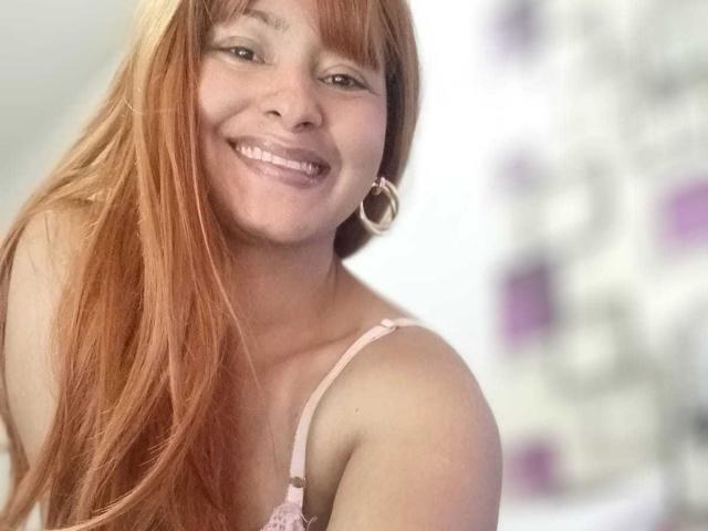 KarlysHoney with long red hair, smiling, wearing pink bra, showing small tits, and large hoop earrings, in a blurry background. She's cute and has a sexy smile.