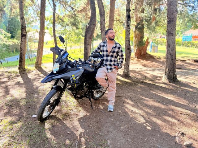 Edahi est appuyé contre une moto noire dans une forêt, portant une chemise à carreaux et un pantalon beige, avec une barbe et des cheveux courts, debout sur de la terre avec des arbres autour..