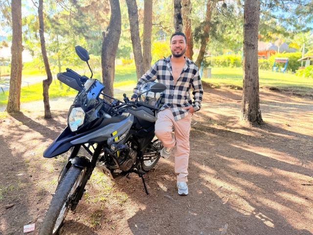 Edahi, con barba y sonriendo, se apoya en una motocicleta negra, vistiendo una camisa a cuadros, pantalones beige y zapatillas blancas, en un bosque iluminado por el sol..