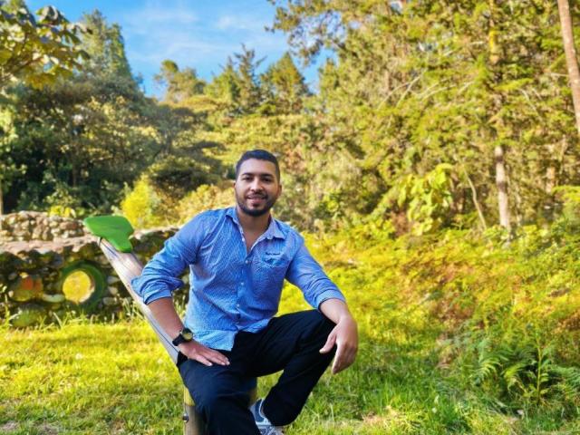 Edahi tiene un trasero atractivo, lleva una camisa azul abotonada y pantalones negros, está haciendo sentadillas en un bosque con hierba, con un tronco de árbol verde al fondo..