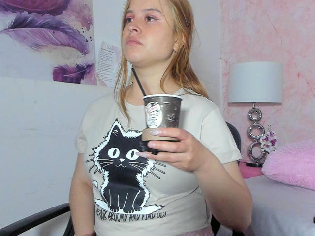 Melani Becker mit blonden Haaren, pinkem Augen-Make-up und einem weißen T-Shirt mit einem schwarzen Katzen-Motiv, die eine Metalltasse mit einem Strohhalm hält und in einem Stuhl sitzt..