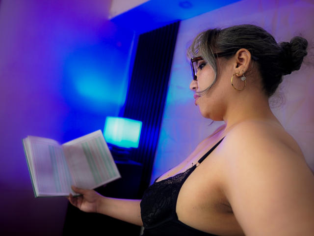 Mariehss with glasses, black lace bra, and hoop earrings, reading a book, showing her tits, in a blue-lit room. Her armpit is visible, hair in a bun.