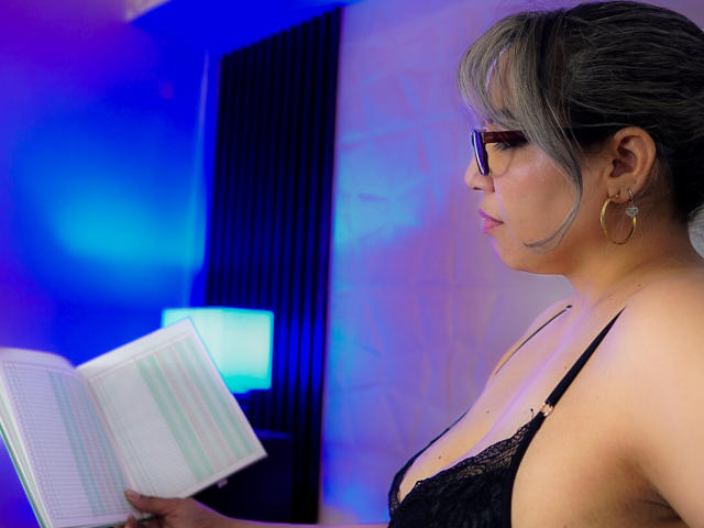 Mariehss with gray hair and glasses, wearing black lace bra, reading a book, blue and purple lighting, hoop earrings, focused expression, holding open book in front of her.