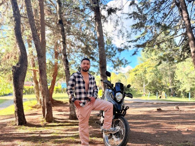 Edahi, con barba y una camisa a cuadros negros y blancos, está de pie con una pierna sobre una motocicleta negra, vistiendo pantalones rosas, en un bosque iluminado por el sol, rodeado de árboles altos..