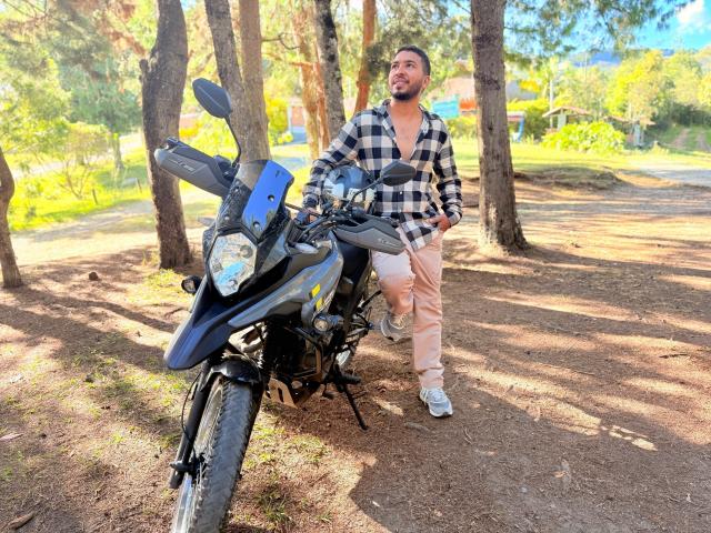 Edahi steht neben einem schwarzen Motorrad mit blauen Akzenten, trägt ein kariertes Hemd und beigefarbene Hosen und blickt leicht lächelnd zur Seite..