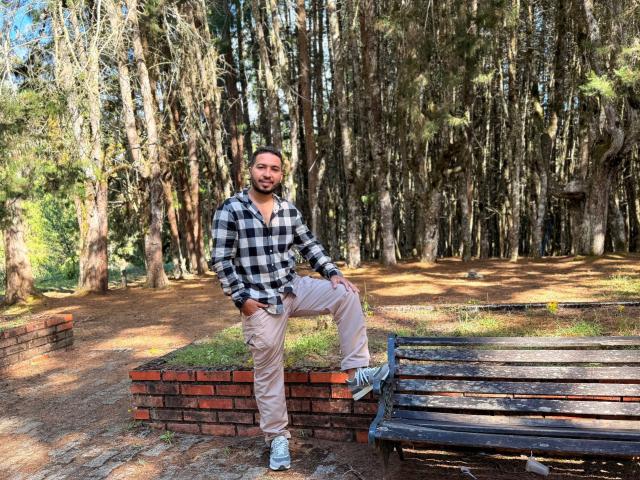 Edahi, em pé com uma perna em um banco de madeira, tem barba, uma faixa de cabeça preta, camisa xadrez, calças bege e tênis brancos. Em primeiro plano, há uma parede de tijolos, e ao fundo, uma área arborizada..